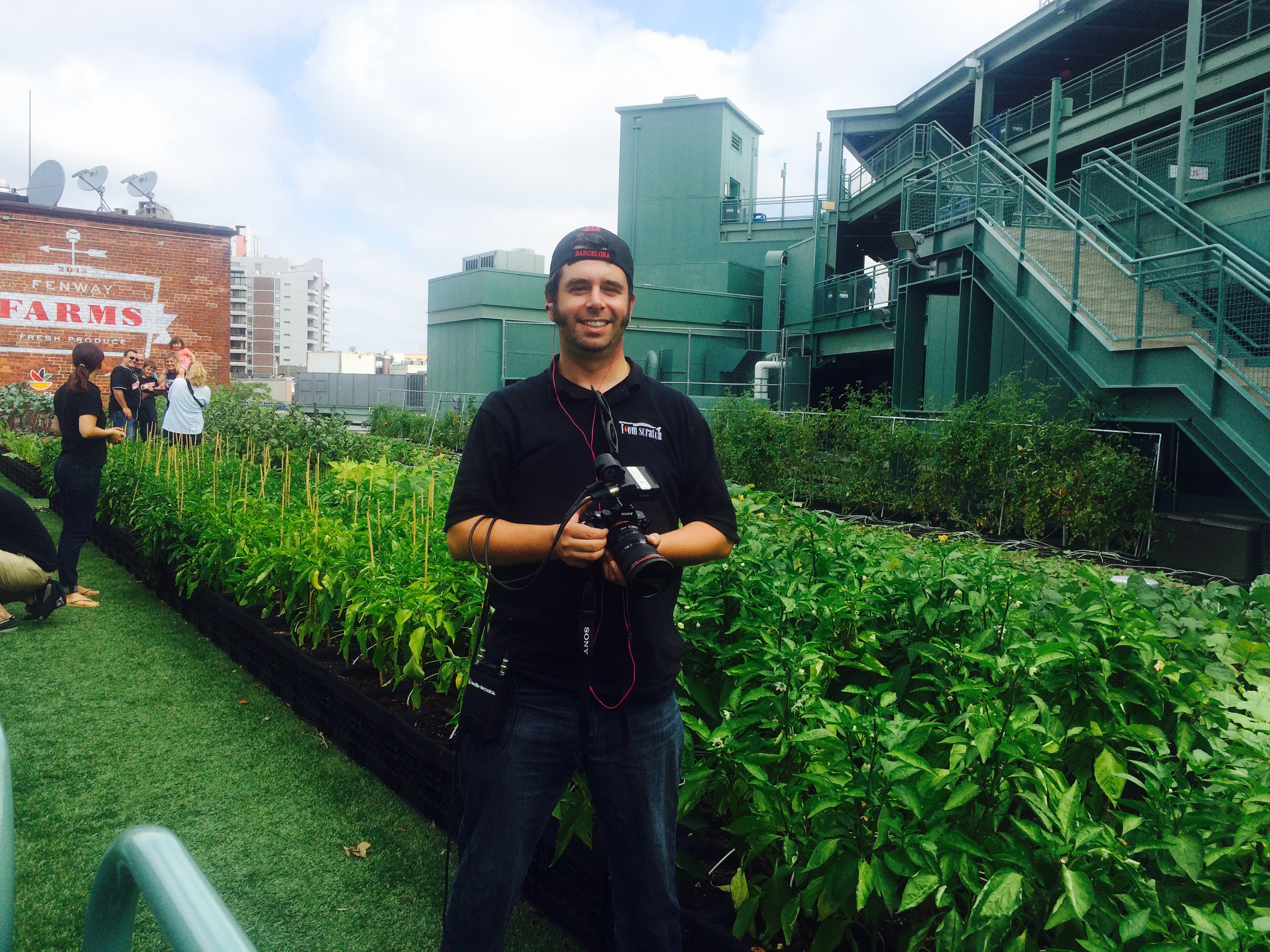 Behind-the-scenes: Ryan holding a camera in a rooftop urban garden at Fenway Park, Boston — green beds, Fenway Farms signage behind.