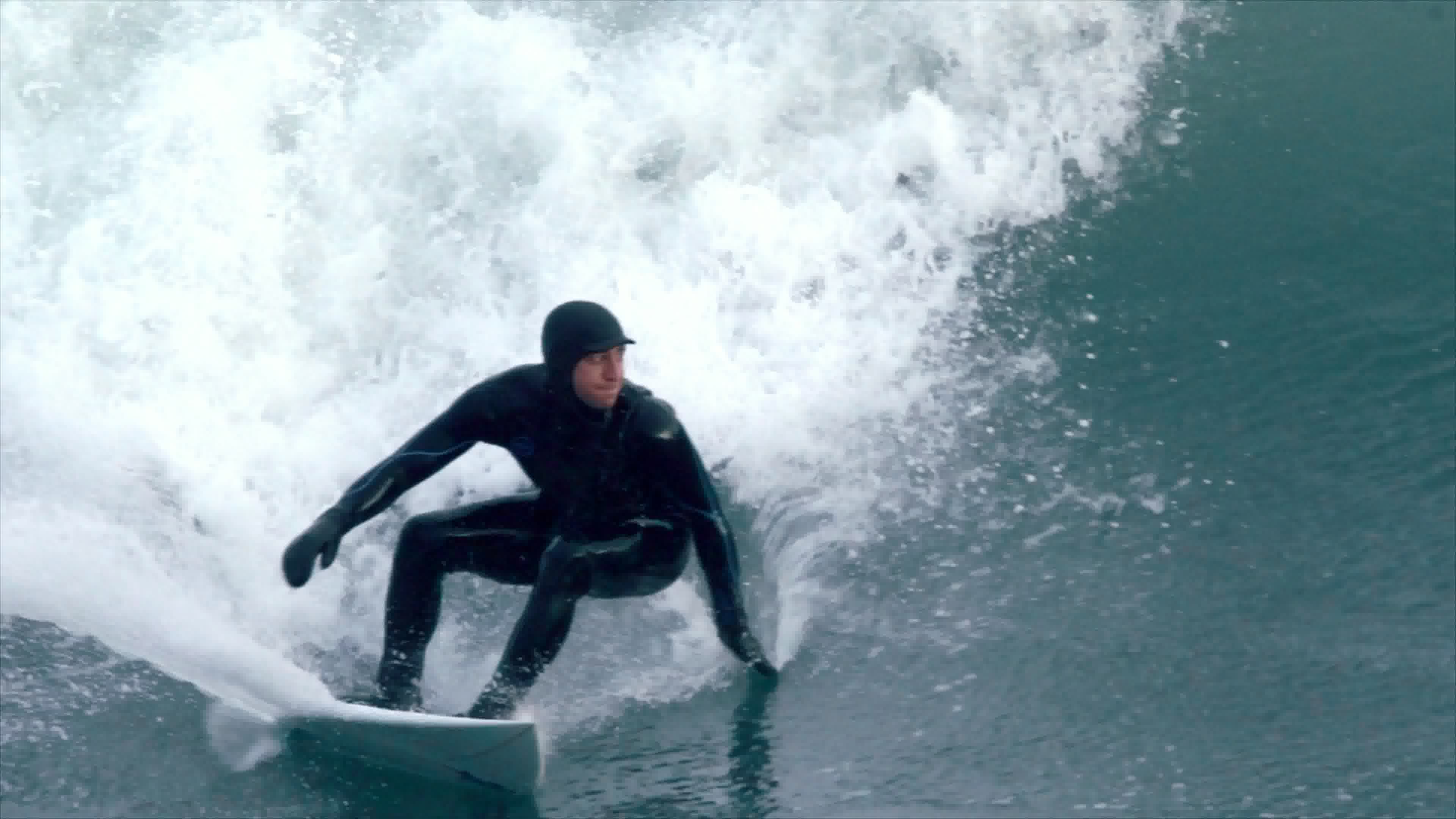 Tasty Waves still — a cold-water surfer in a hooded wetsuit carving the bottom of a gray-green wave, whitewash exploding behind.